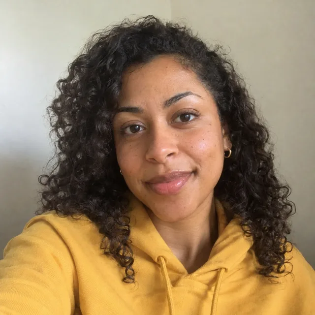 Phone selfie of a fictional creator with curly hair and a yellow hoodie, used as the input photo